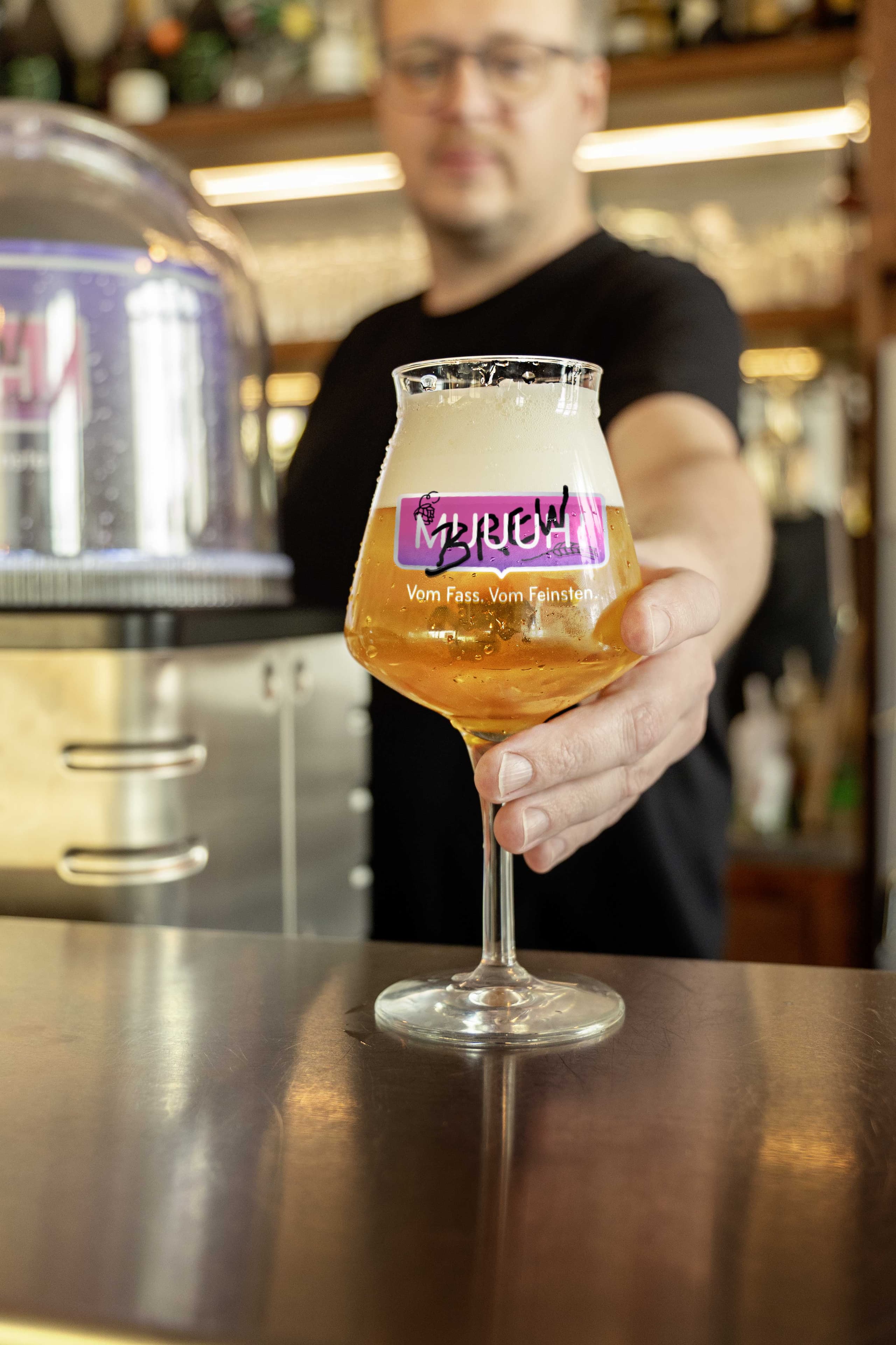 Craft-Beer im Glas mit MUUUH!-Logo und Claim 'Vom Fass. Vom Feinsten.' vor Brauerei-Kulisse