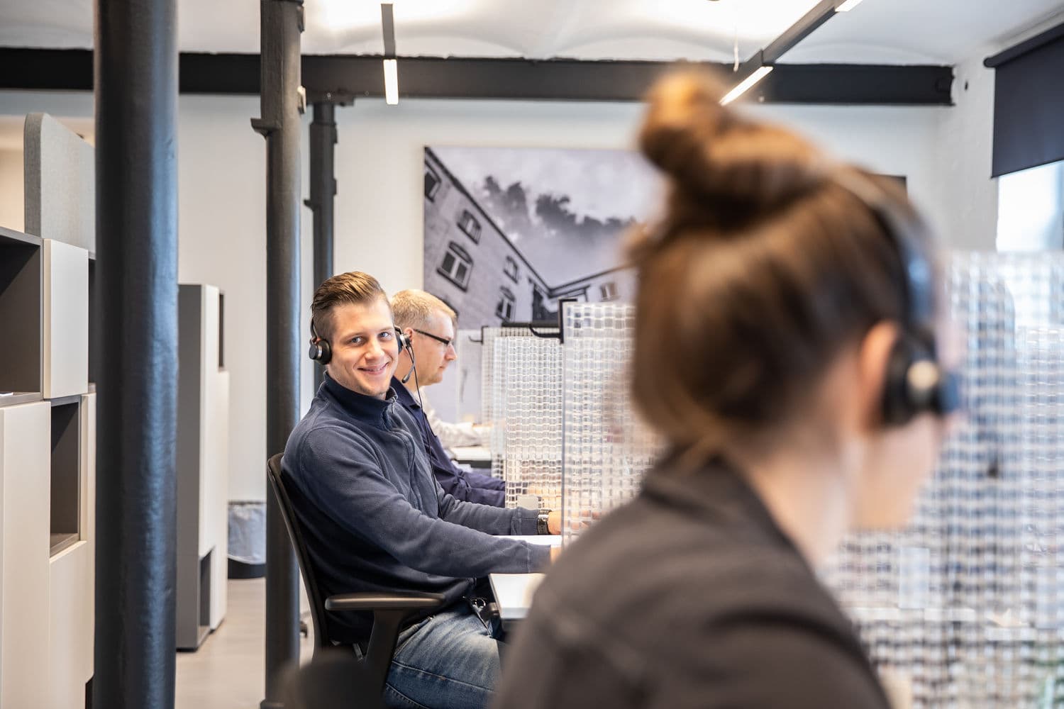 Lächelnder Kundenberater mit Headset im Callcenter, im Vordergrund eine Kollegin mit Headset