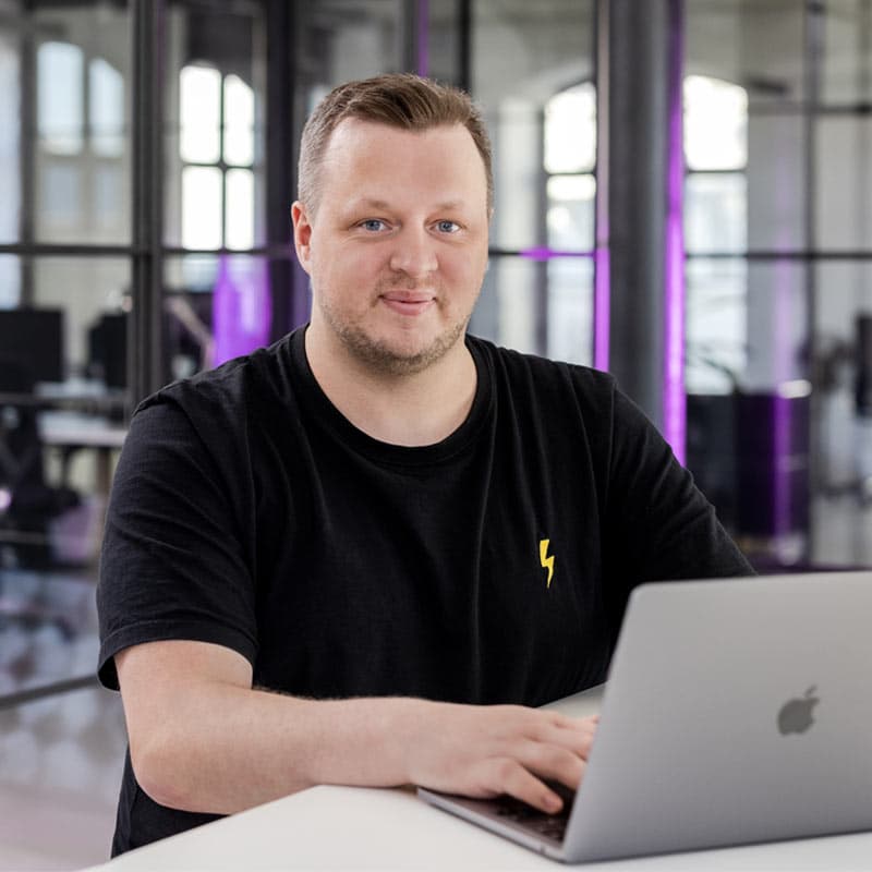 John, lächelnd vor einem Laptop in einem modernen Büro, schwarzes T-Shirt mit gelbem Blitz-Logo