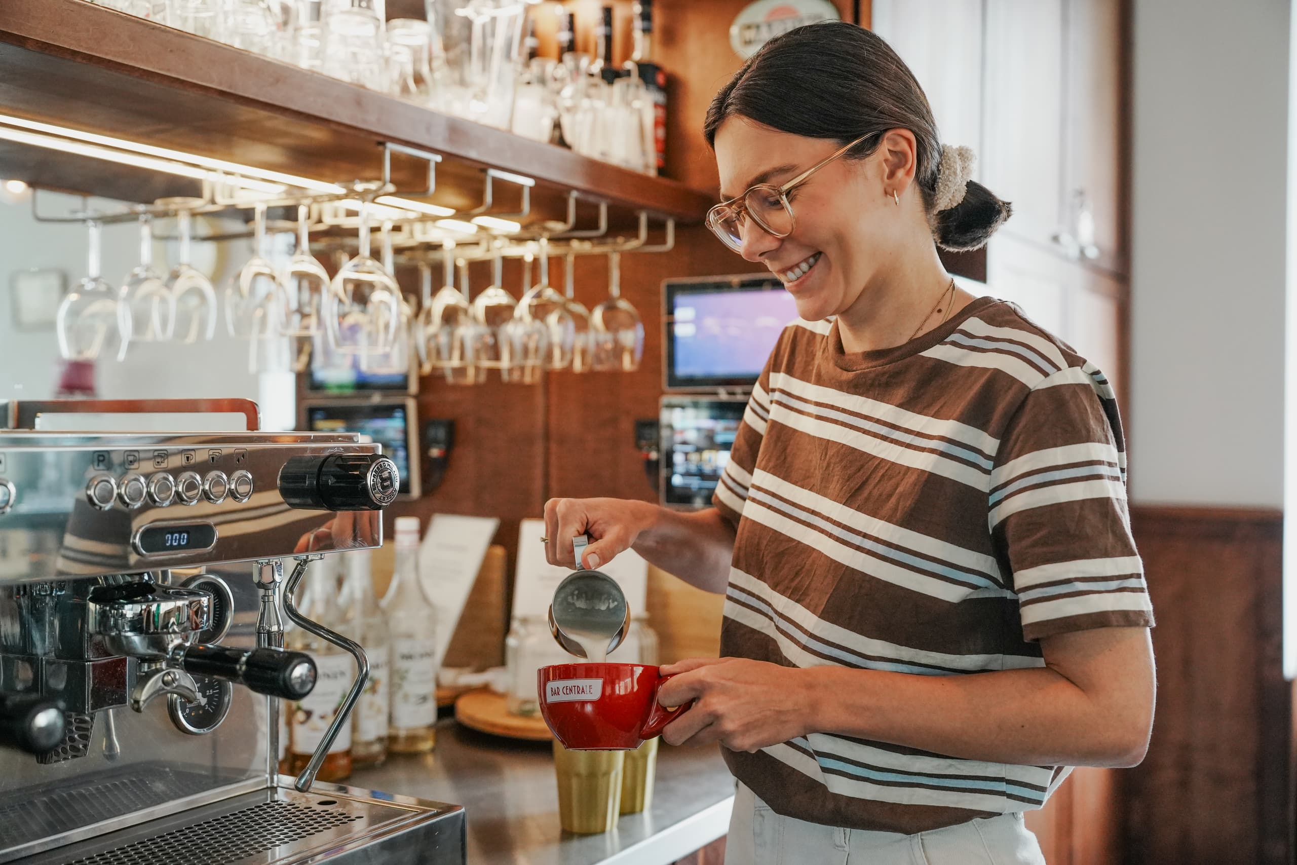 Lächelnde Mitarbeiterin bereitet an einer Espressomaschine einen Kaffee zu und schäumt Milch auf