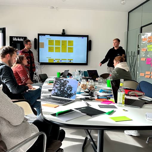 Workshop-Gruppe beim Kickoff mit MUUUH! und Seedhouse: Teilnehmende sitzen am Tisch, ein Moderator präsentiert Wireframes auf einem großen Bildschirm