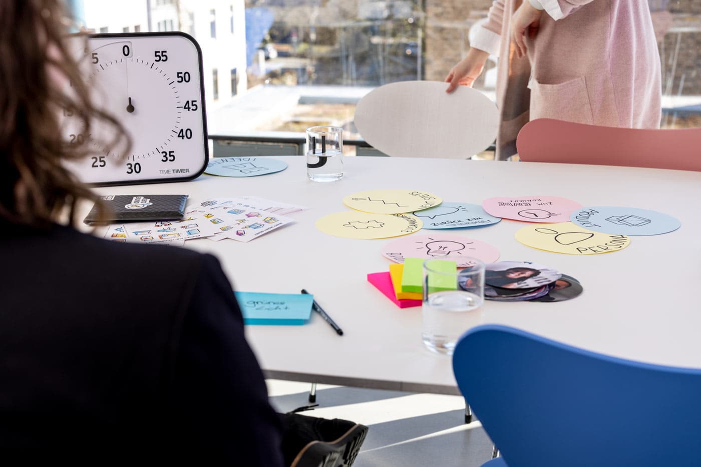 Workshop-Tisch mit bunten Moderationskarten, Post-its und einer analogen Timer-Uhr – zwei Personen arbeiten gemeinsam