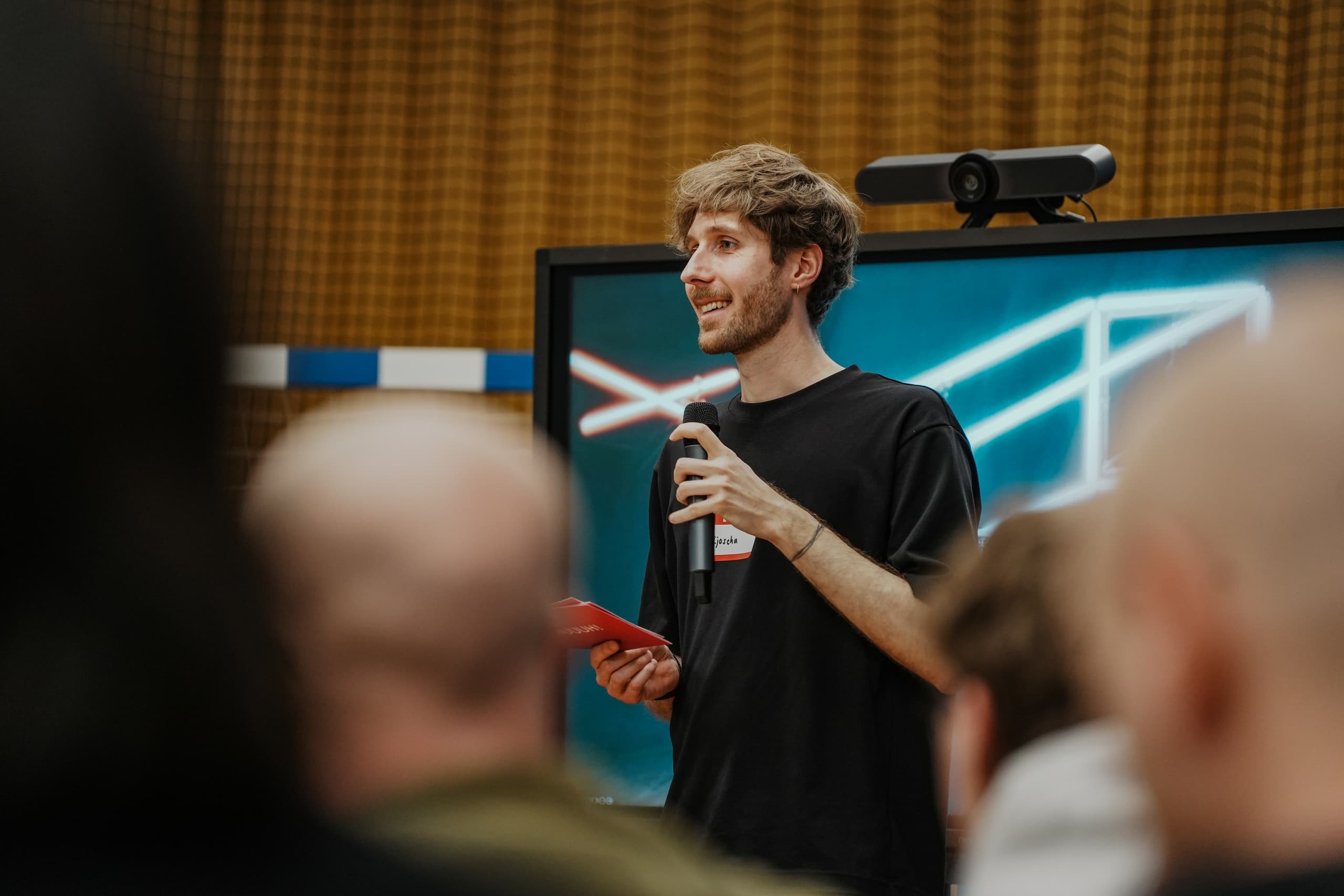 Junger Mann mit Mikrofon hält eine Präsentation vor Publikum, im Hintergrund ein Bildschirm mit Grafik