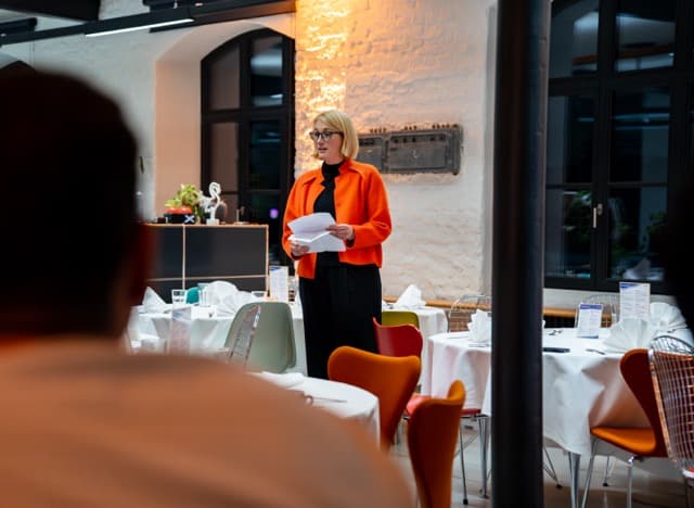 Eine Frau in orangefarbener Jacke steht mit Unterlagen in der Hand vor einem Publikum in einem stilvollen Restaurant