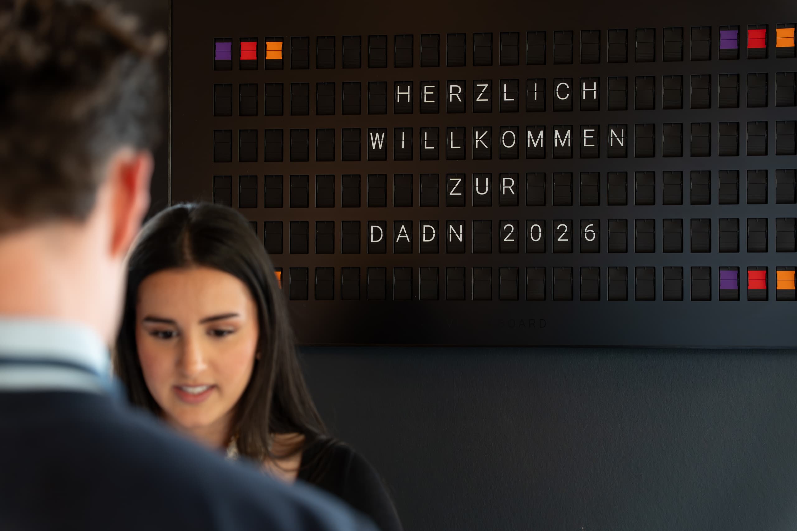 Anzeigetafel mit der Aufschrift 'HERZLICH WILLKOMMEN ZUR DADN 2026', im Vordergrund zwei Personen unscharf
