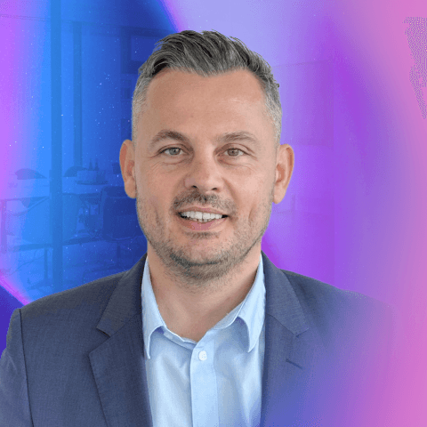 Genc Rama, lächelnd im dunkelblauen Sakko, vor violett-blauem Hintergrund