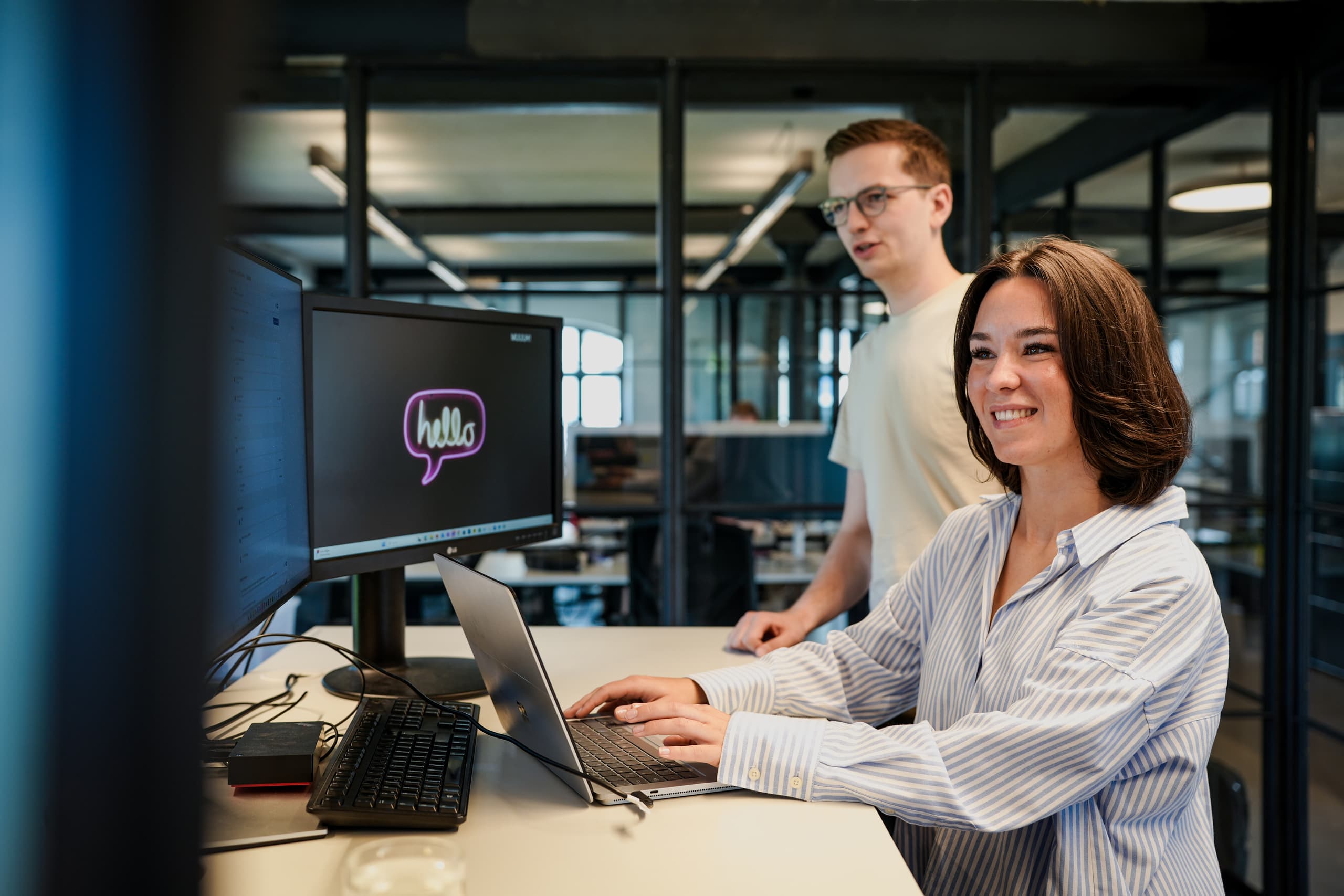 Zwei junge Mitarbeitende arbeiten lächelnd an Laptop und Desktop-Monitor mit MUUUH-Logo im Büro