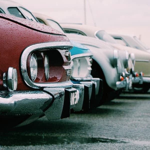 Mehrere geparkte Oldtimer in einer Reihe, Nahaufnahme der Frontpartien mit Chromstoßstangen bei regnerischem Wetter