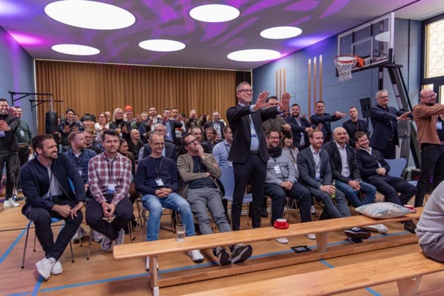 Große Gruppe von Teilnehmenden beim MUUUH!nwalk 2025 in einer Sporthalle, ein Redner hebt die Hände