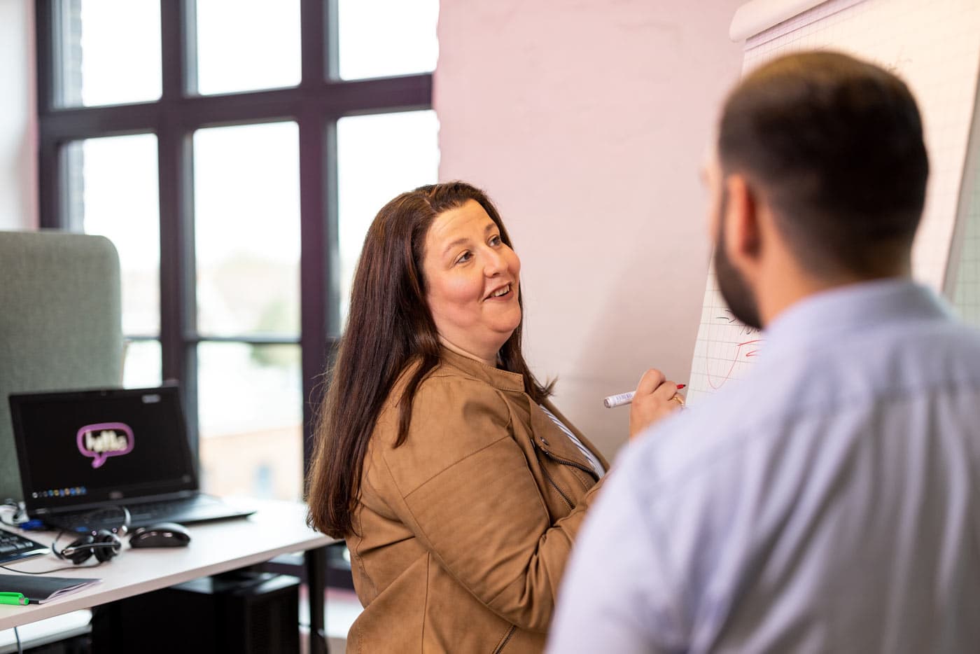 Maria erklärt einem Kollegen etwas am Whiteboard und lächelt ihn dabei an