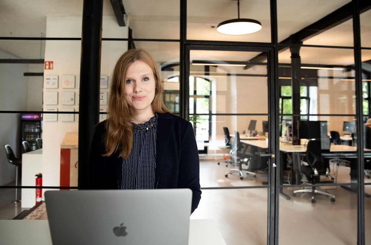 Madlen Hund steht vor einem Laptop in einem modernen Büro mit Glaswänden und offener Arbeitsumgebung