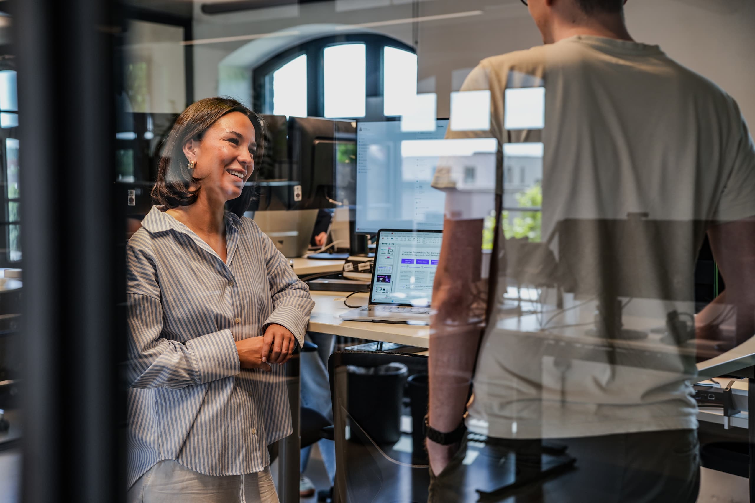 Leoni Hock lächelt im Gespräch mit einem Kollegen in einem modernen Büro mit Computerarbeitsplätzen im Hintergrund