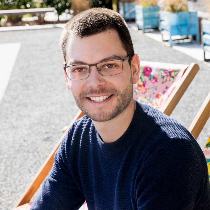 Frank, lächelnd im Porträt, trägt eine Brille und einen dunkelblauen Pullover, im Hintergrund ein Liegestuhl