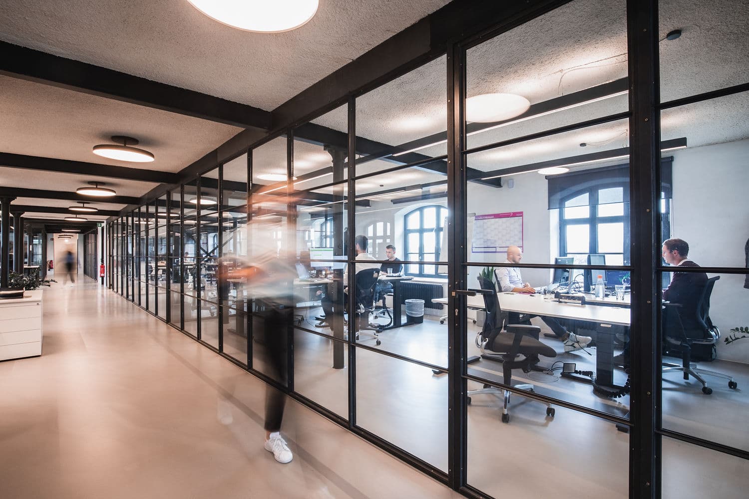 Modernes Büro mit Glastrennwänden: Mitarbeitende arbeiten konzentriert im Team, eine Person läuft dynamisch am Gang vorbei