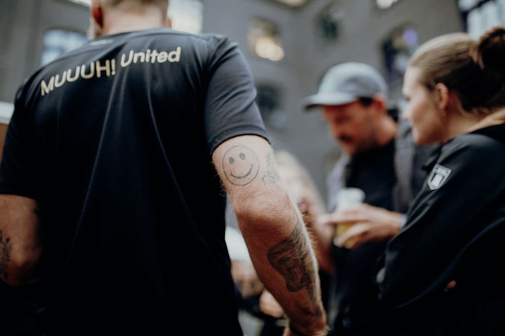 Person im MUUUH! United T-Shirt mit Smiley-Tattoo am Unterarm, im Hintergrund weitere Personen bei einer Veranstaltung