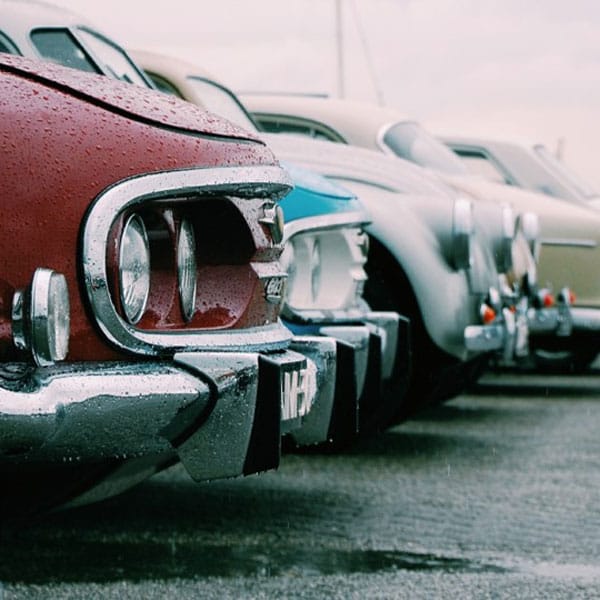 Mehrere geparkte Oldtimer in einer Reihe, Nahaufnahme der Frontpartien mit Chromstoßstangen bei regnerischem Wetter