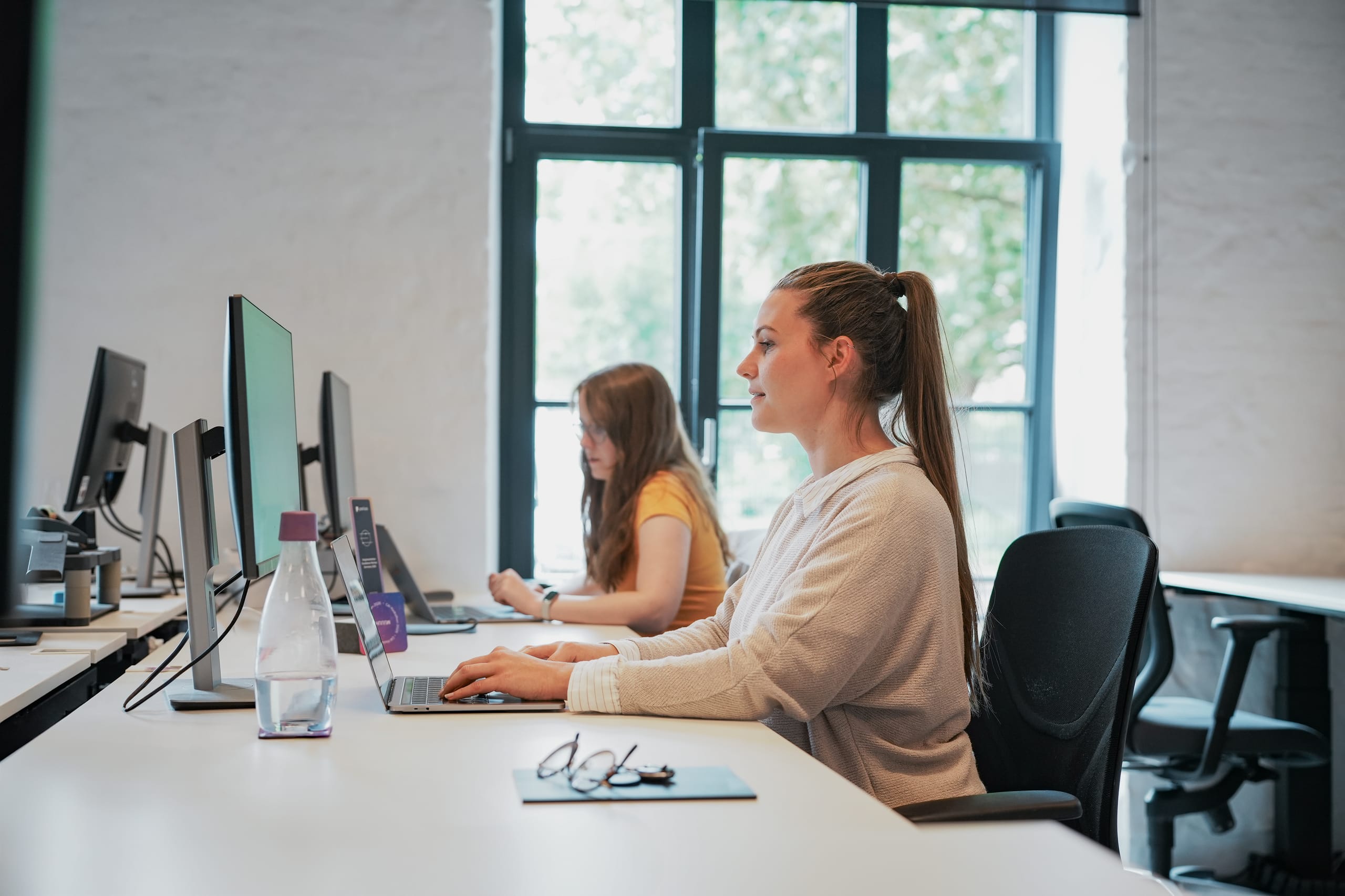 Mitarbeiterin arbeitet konzentriert am Laptop in einem modernen Büro – im Hintergrund eine weitere Person am Computer