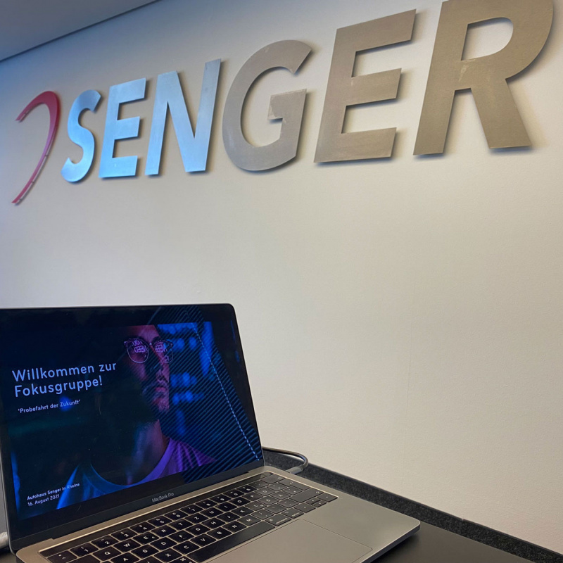Das Logo der Unternehmensgruppe hängt an einer Wand, davor steht ein Macbook mit einer geöffneten Präsentation